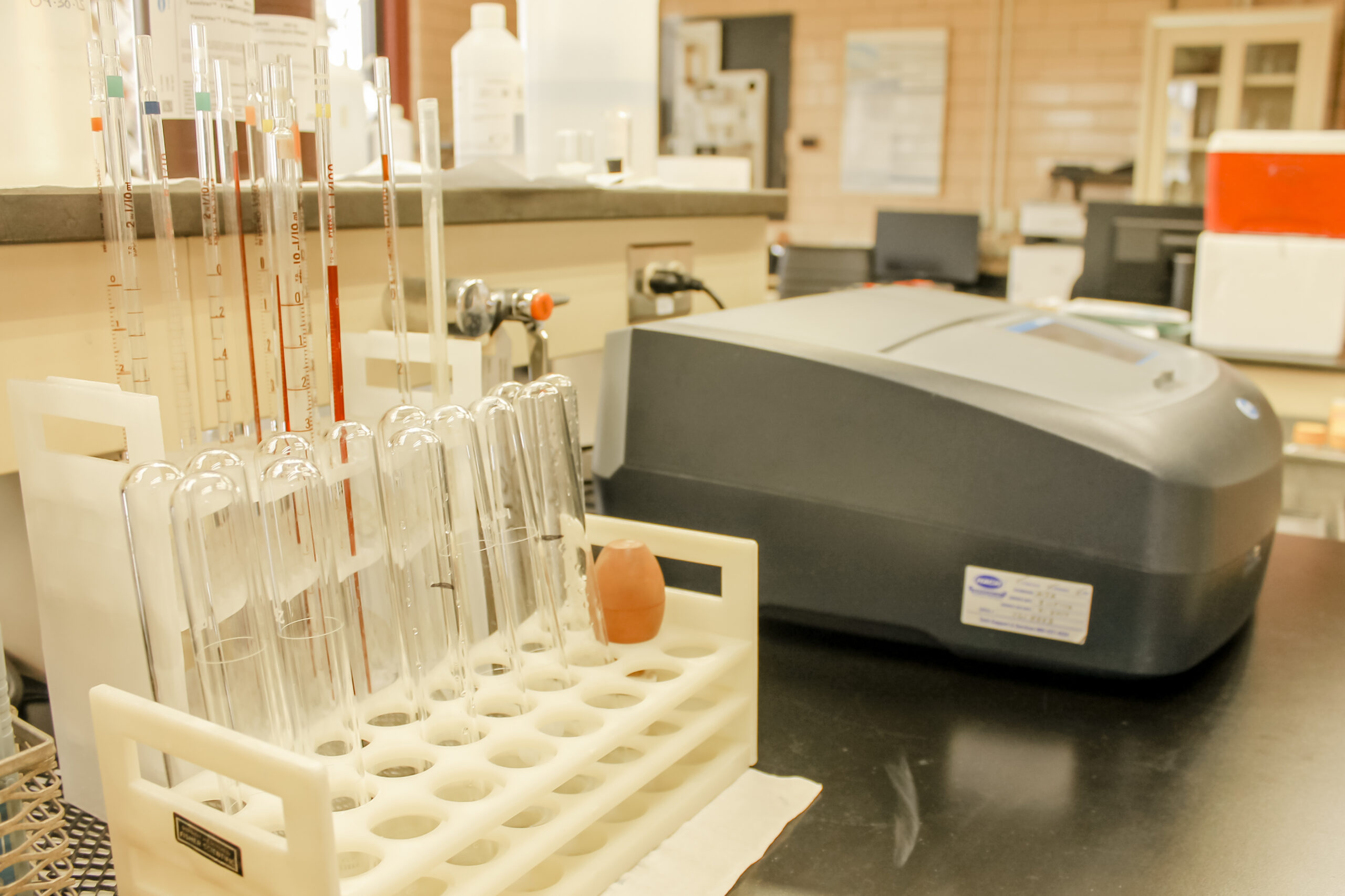Scientist in lab coat and gloves examines clear liquid in Erlenmeyer flask, wearing safety goggles.