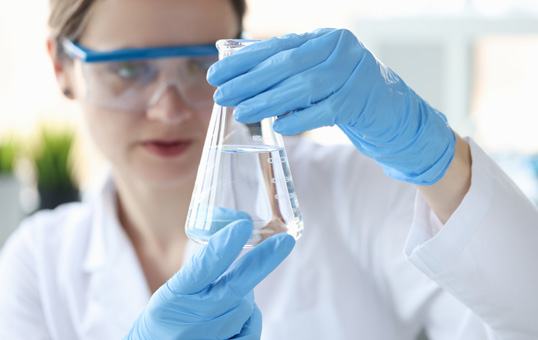 Scientist in lab coat and gloves examines clear liquid in Erlenmeyer flask, wearing safety goggles.