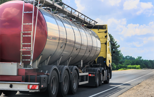 Tanker truck transporting liquid on a highway under a partly cloudy sky. The truck is yellow and silver.