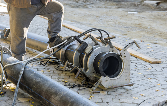 Worker using fusion welding machine to join large black plastic pipes on a cobblestone surface.