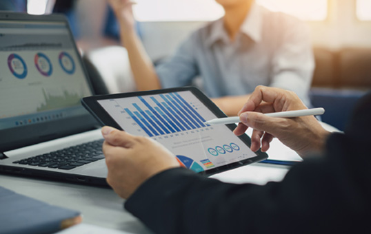 Business professional analyzing charts and graphs on a tablet during a meeting, with a laptop also displaying data.
