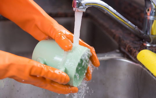 Hands in orange gloves washing a green mug under running water in a stainless steel sink.