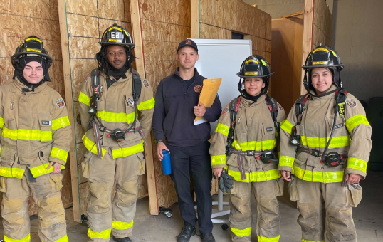 Group of firefighters in gear with instructor holding paperwork, posing indoors.
