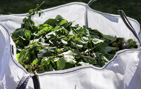 White bulk bag filled with green leaves and small branches, ready for yard waste disposal.