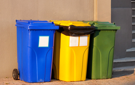 Three wheeled trash bins: blue, yellow, and green, lined up against a wall for waste sorting and recycling.