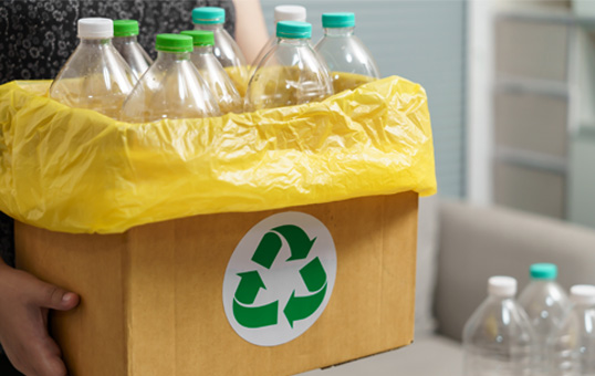 Person holding a cardboard box filled with empty plastic bottles for recycling, marked with a green recycling symbol.