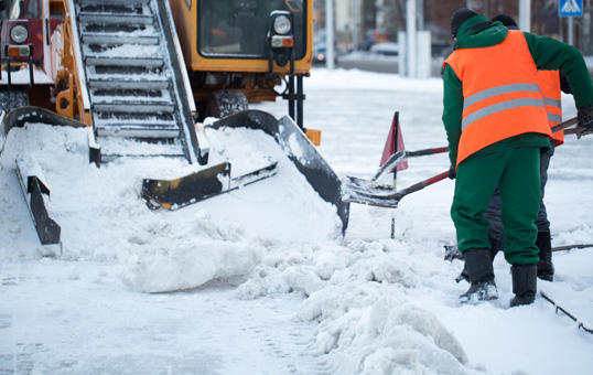 Workers in orange vests shovel snow next to a snowplow on a city street after a snowfall.