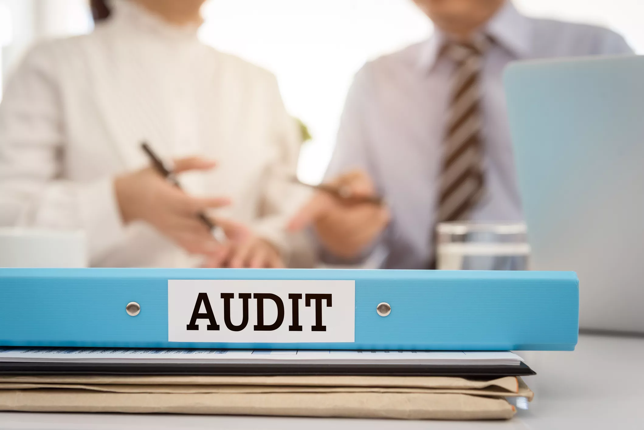 Blue audit binder on a desk with two people blurred in the background, suggesting a business meeting or audit review.