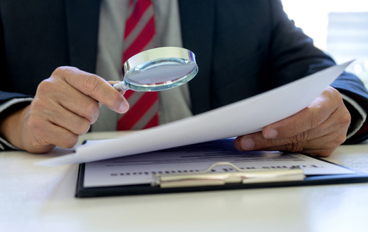 Man in suit examines document with magnifying glass on a clipboard. Focus on review and attention to detail.