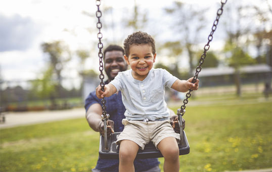 Smiling toddler enjoys a swing at the park, pushed by a smiling adult.