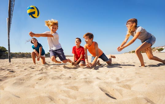 Children playing volleyball on a sunny beach, one boy jumping to hit the ball over the net.