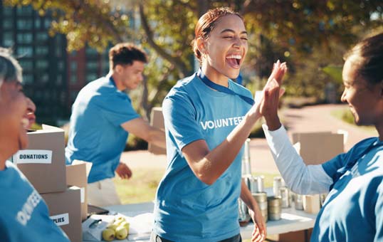 Volunteers in blue shirts give a high five at an outdoor charity event with donation boxes and canned goods.