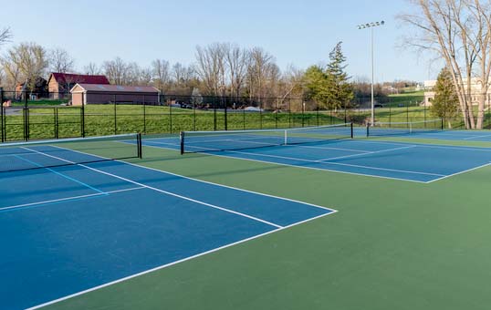 Two blue and green outdoor tennis courts with nets, surrounded by trees and a fence on a sunny day.