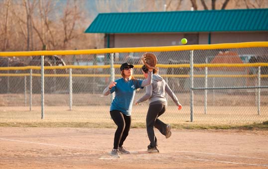 Two women playing catch with a softball on a sunny day at a park.