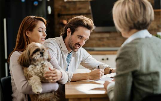 Couple signs paperwork at table with advisor, dog on lap. Focus on signing. Represents financial planning or home buying.