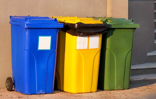 Three wheeled trash cans: blue, yellow, and green, standing side-by-side against a wall.