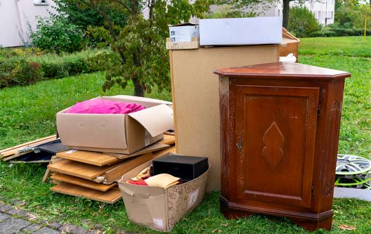 Pile of unwanted household items on grass, including boxes, furniture, and miscellaneous objects, likely for disposal.