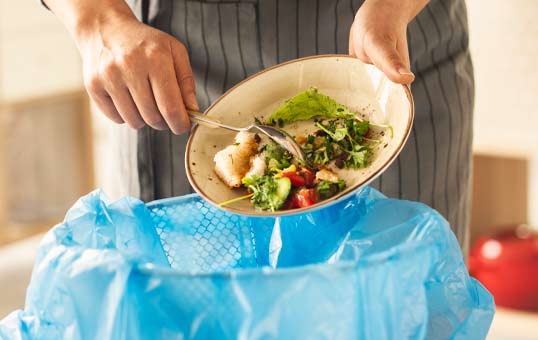 Person discarding leftover food into a blue trash bag, emphasizing food waste reduction.