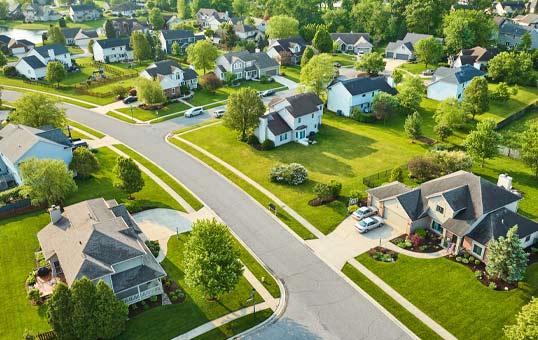Aerial view of a suburban neighborhood with houses, green lawns, and tree-lined streets on a sunny day.