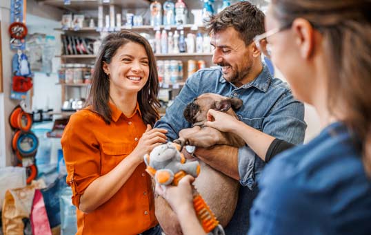 Couple smiling while holding a puppy in a pet store, with toys and supplies visible in the background.
