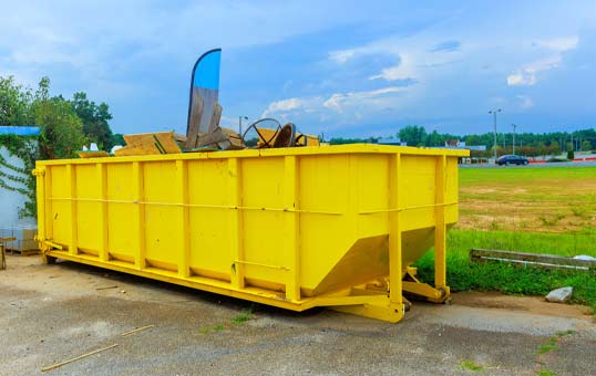 Bright yellow roll-off dumpster overflowing with construction debris, parked on a concrete surface outdoors.