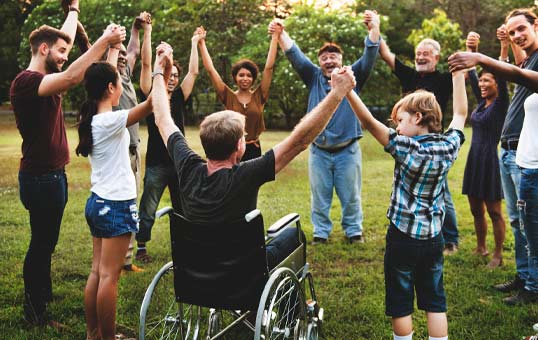 Diverse group of people holding hands in a circle outdoors, including a person in a wheelchair.