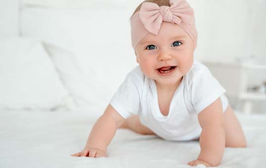 Baby in white onesie and pink bow crawling on white bed, smiling at the camera.