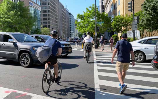 City street scene with cyclists, pedestrians crossing, and cars. Daytime, sunny weather, urban environment.