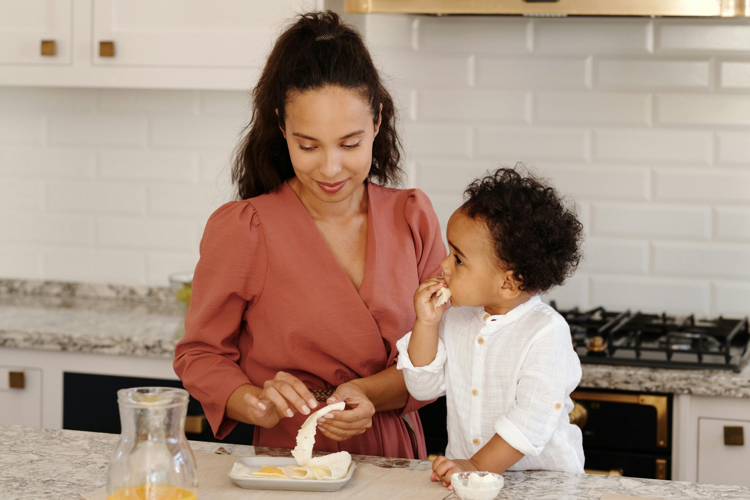 Mother peeling an orange for her toddler in a bright, modern kitchen. The child is eating a slice.