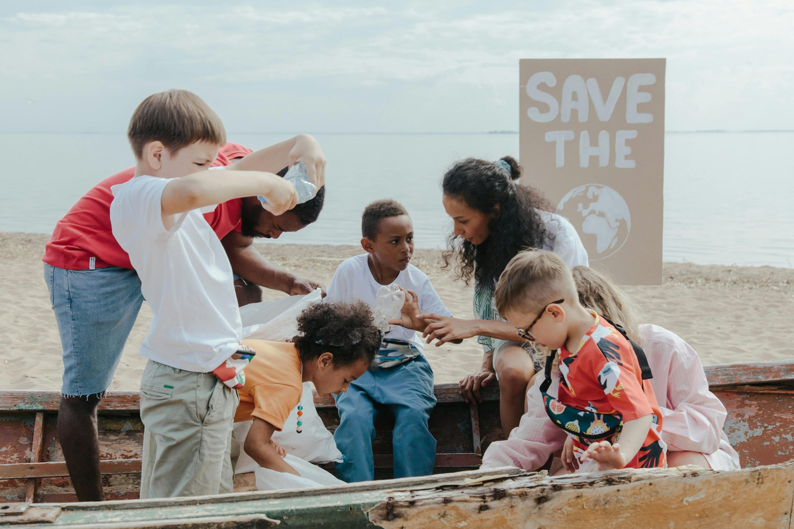 Group of children and adults cleaning up trash on a beach near a boat, with a 'Save the' sign in the background.