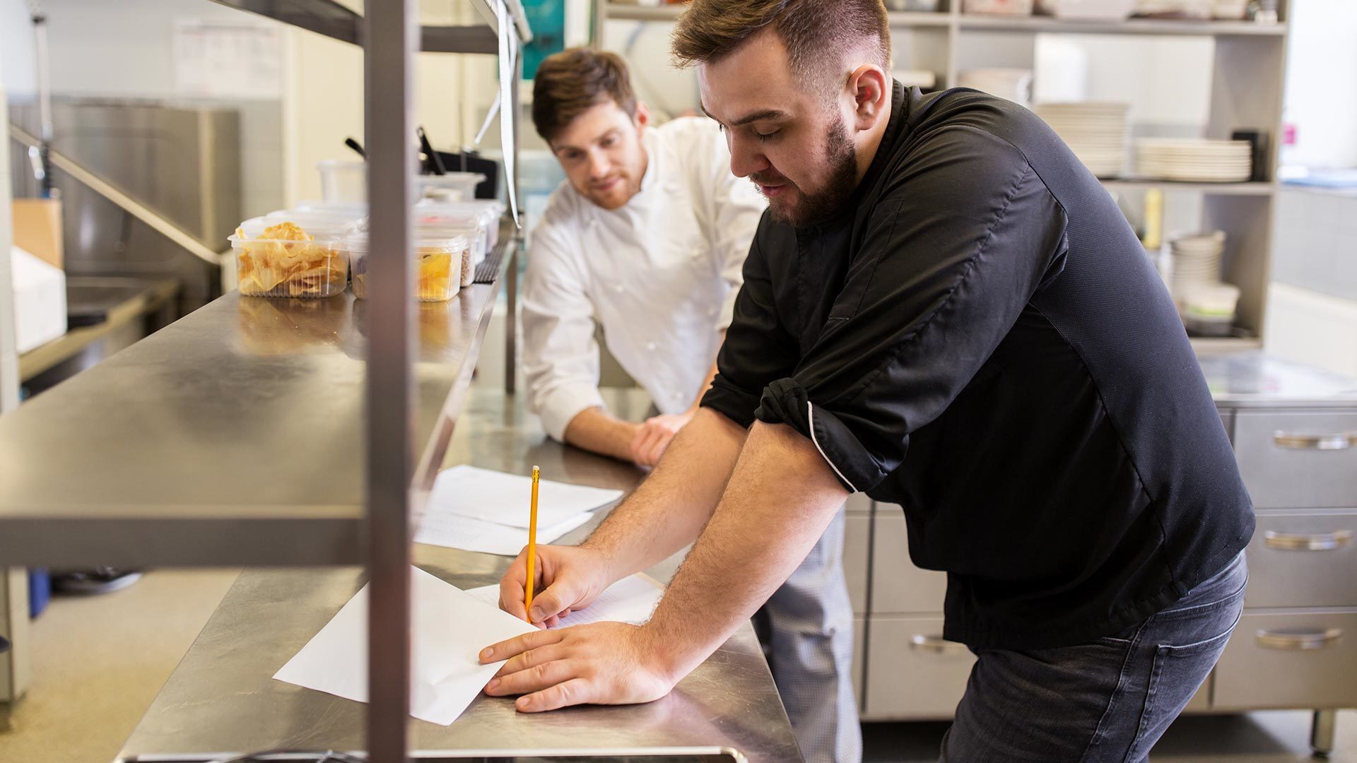Two chefs reviewing paperwork on a stainless steel counter in a commercial kitchen.