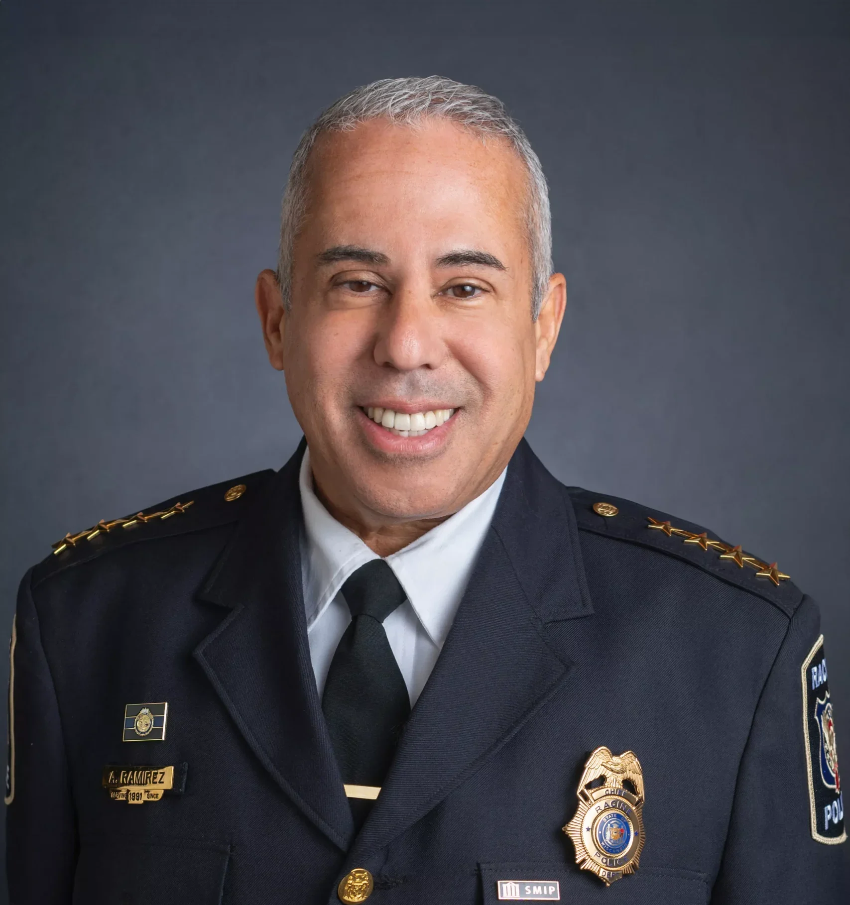 Official portrait of a police chief in uniform, smiling against a dark gray background.