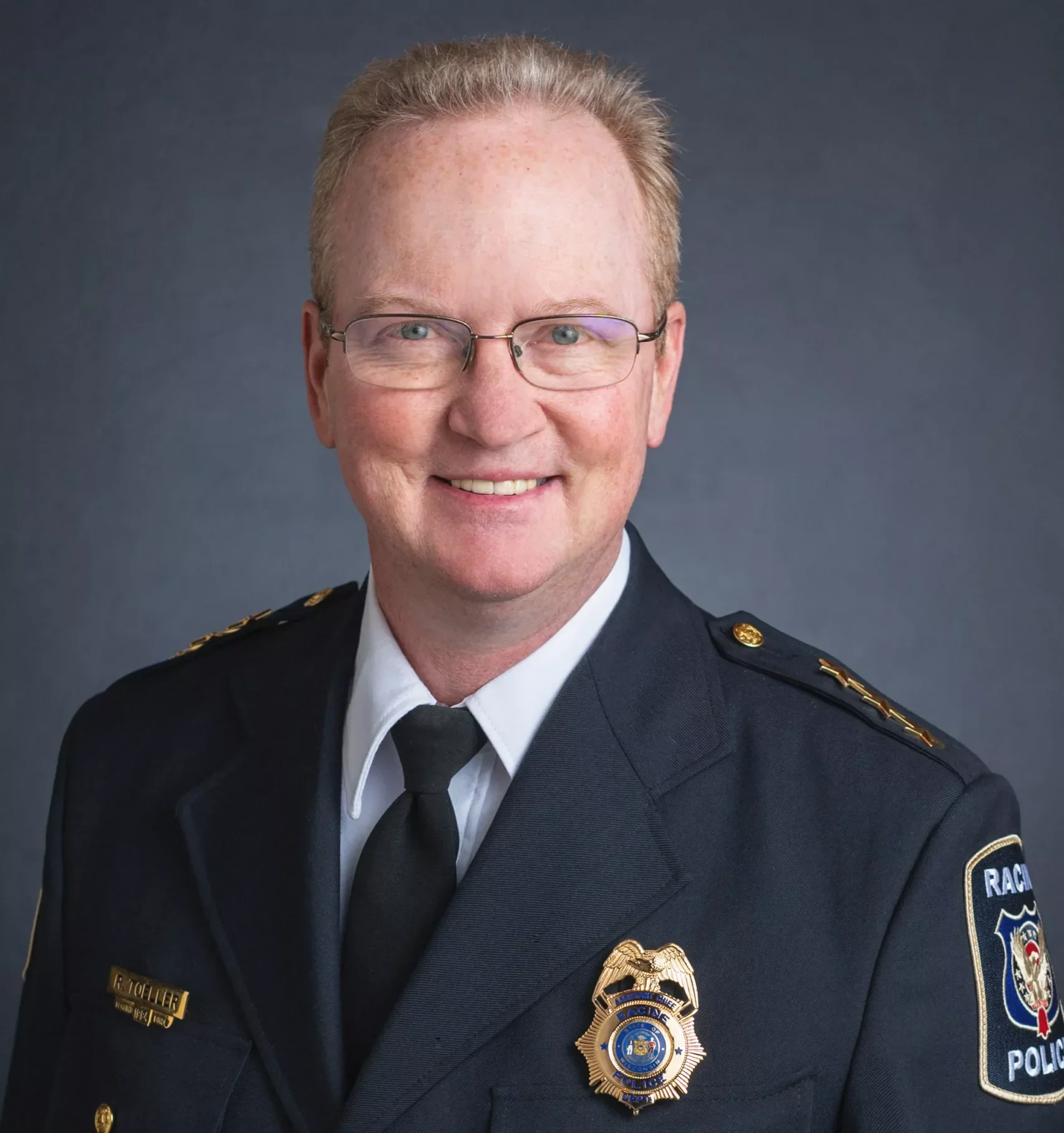 Headshot of a smiling police chief in uniform with glasses against a gray background.