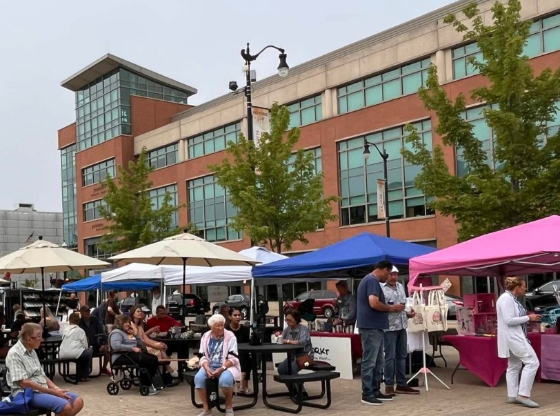 Outdoor market scene with vendor tents and people browsing in front of a brick building on a cloudy day.
