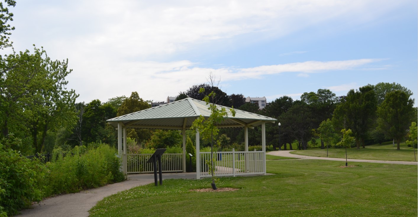 Gazebo in a park setting with a winding path, green grass, and trees under a partly cloudy sky.