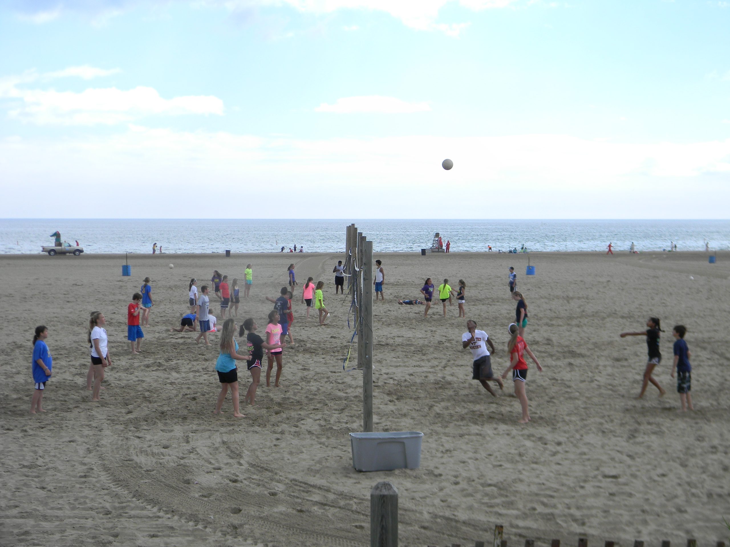 Group playing volleyball on a sandy beach with ocean and sky in background. Volleyball is in the air.