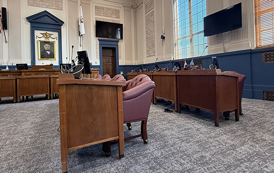 Empty government chamber with wooden desks, leather chairs, and a large window. A screen is mounted on the wall.