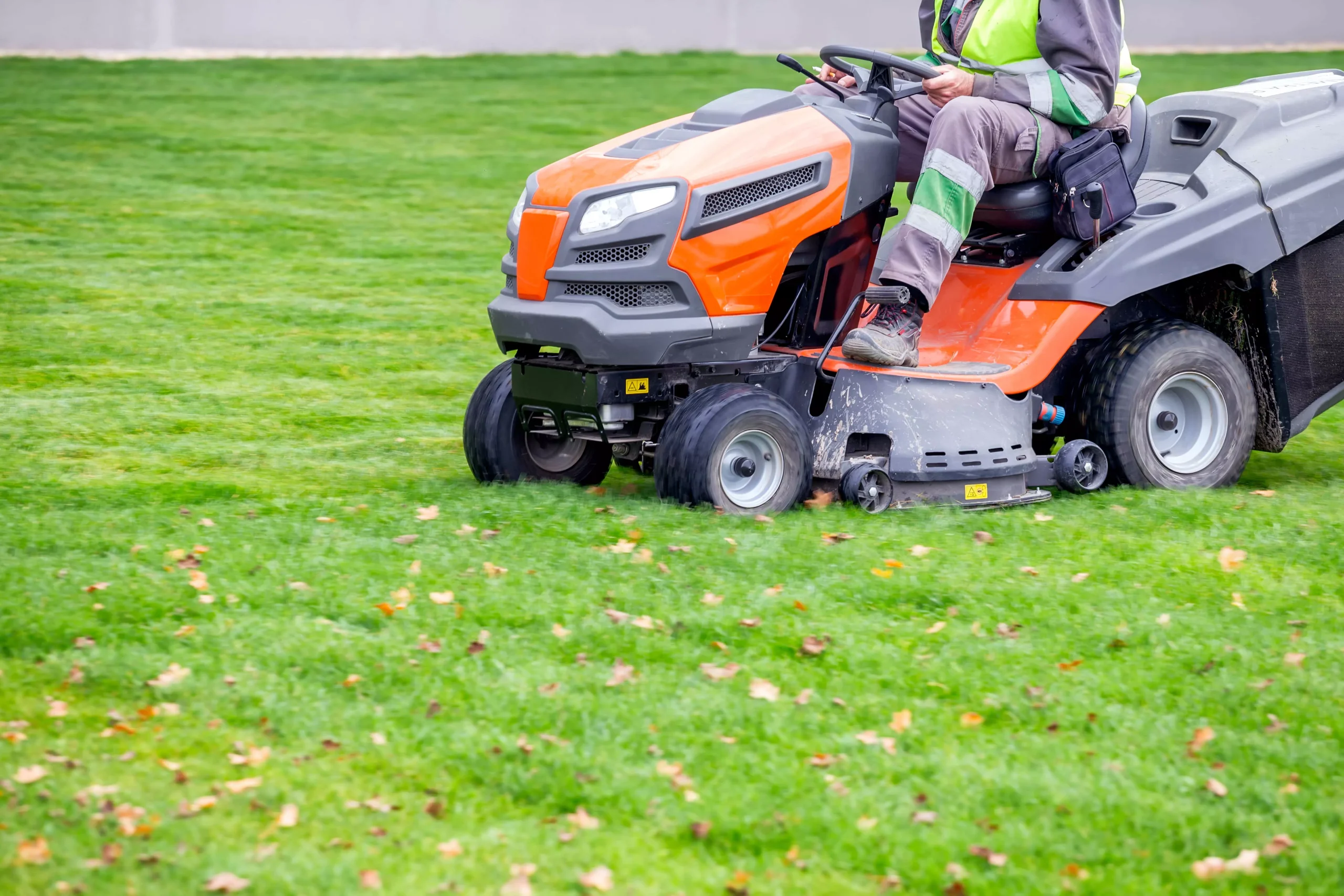 Person mowing a green lawn with an orange and gray riding lawnmower. Grass clippings are visible.