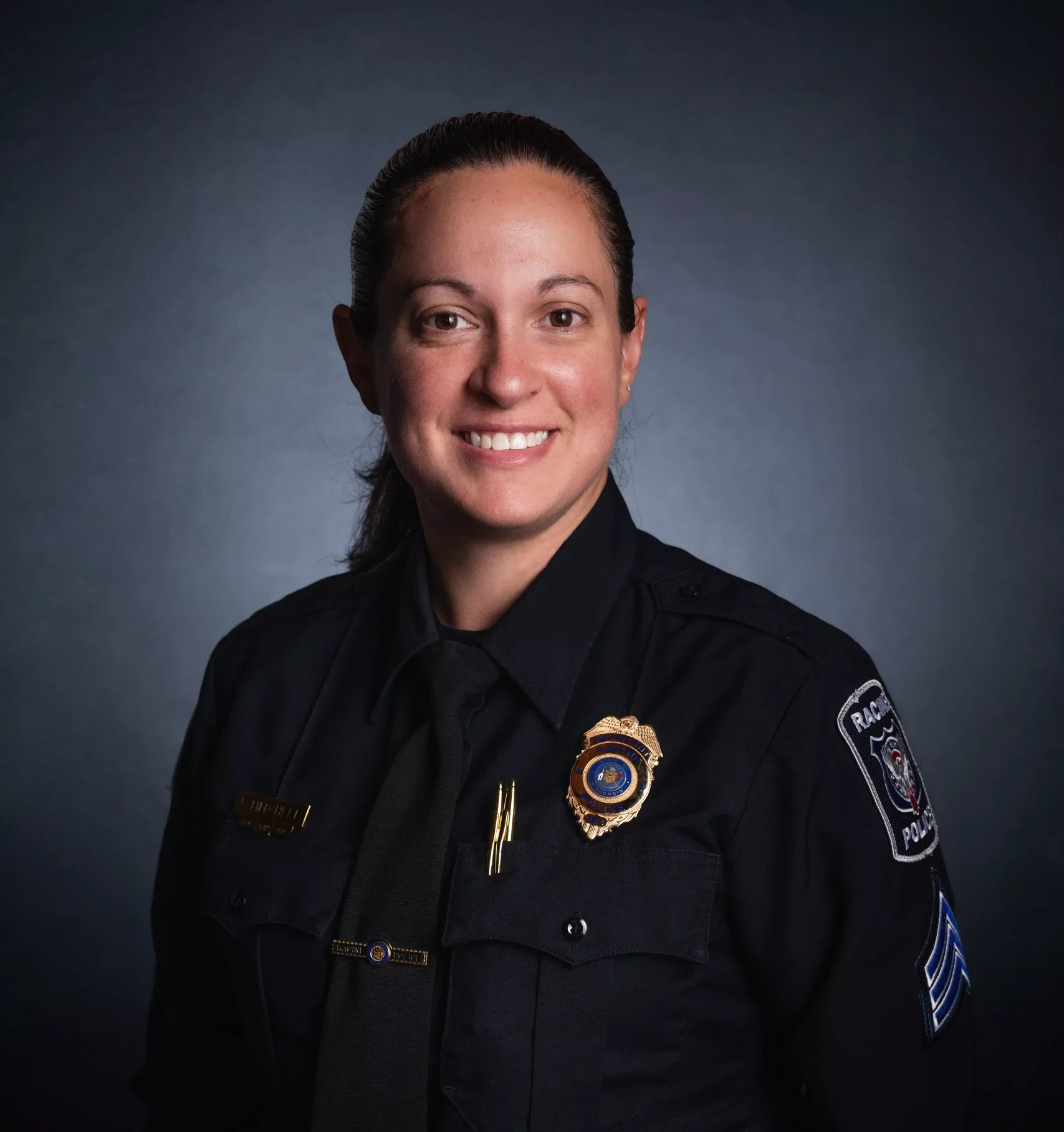 Headshot of a smiling female police officer in uniform against a dark background.