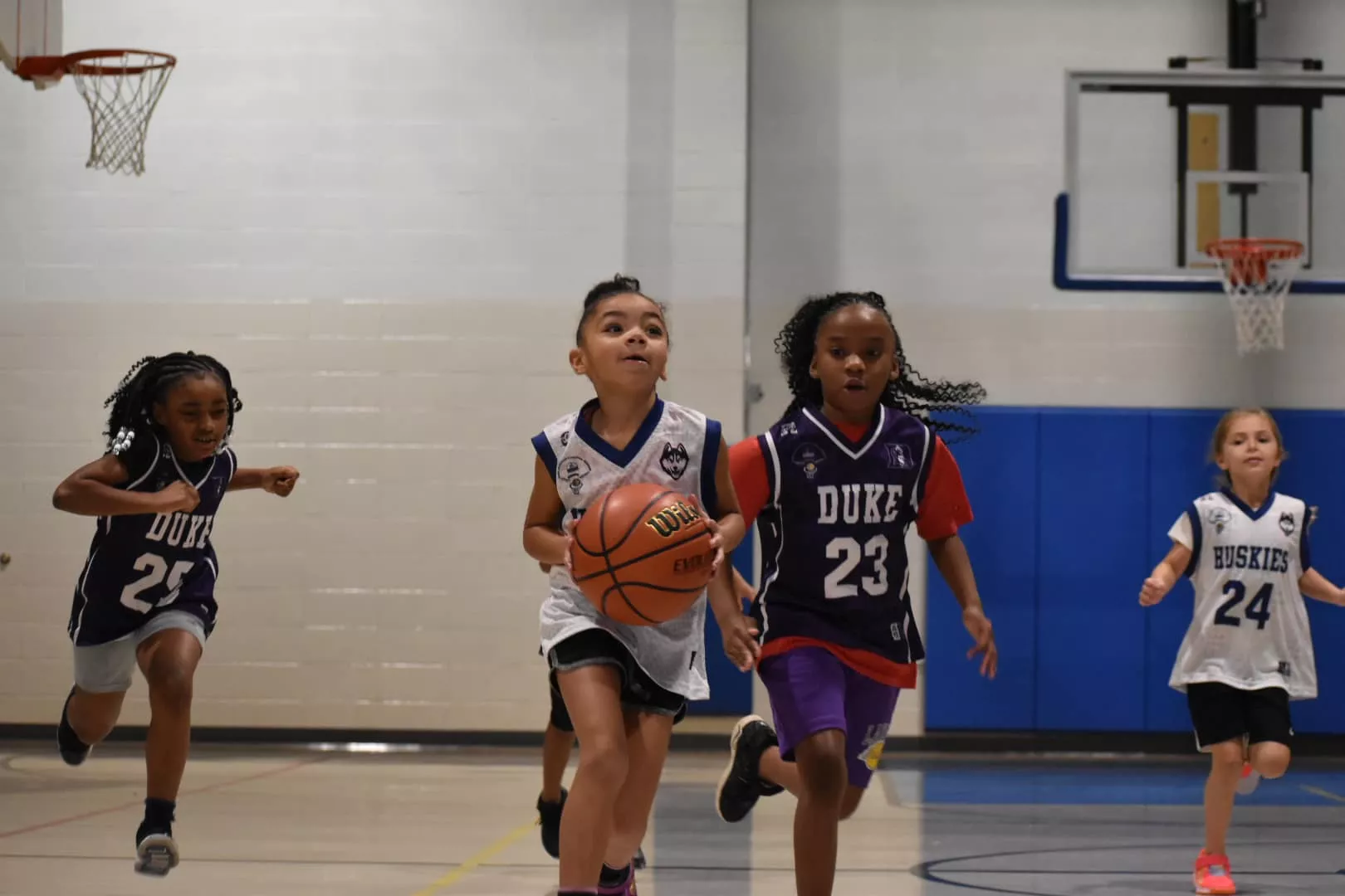 Children in basketball jerseys run down a court during a youth basketball game, focused on the ball.