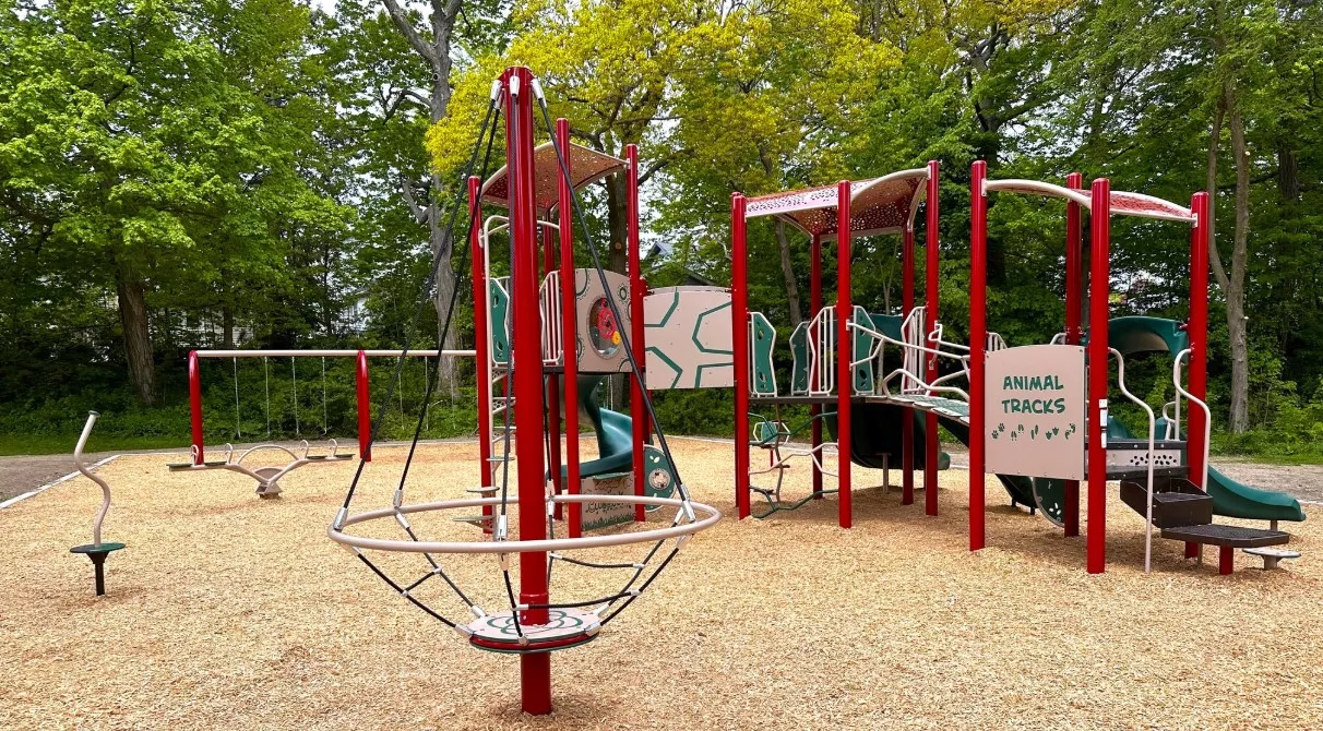 Playground with red and white equipment, including a merry-go-round, slide, and climbing bars, surrounded by wood chips.