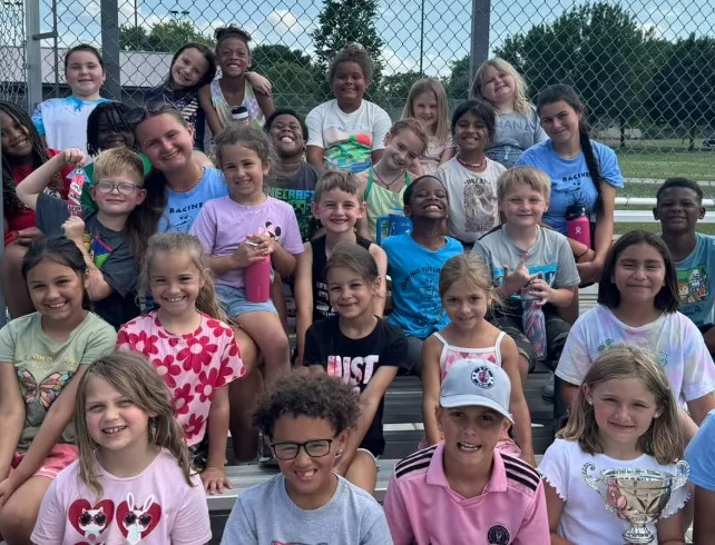 Group of smiling elementary school students and teacher posing on bleachers outdoors.