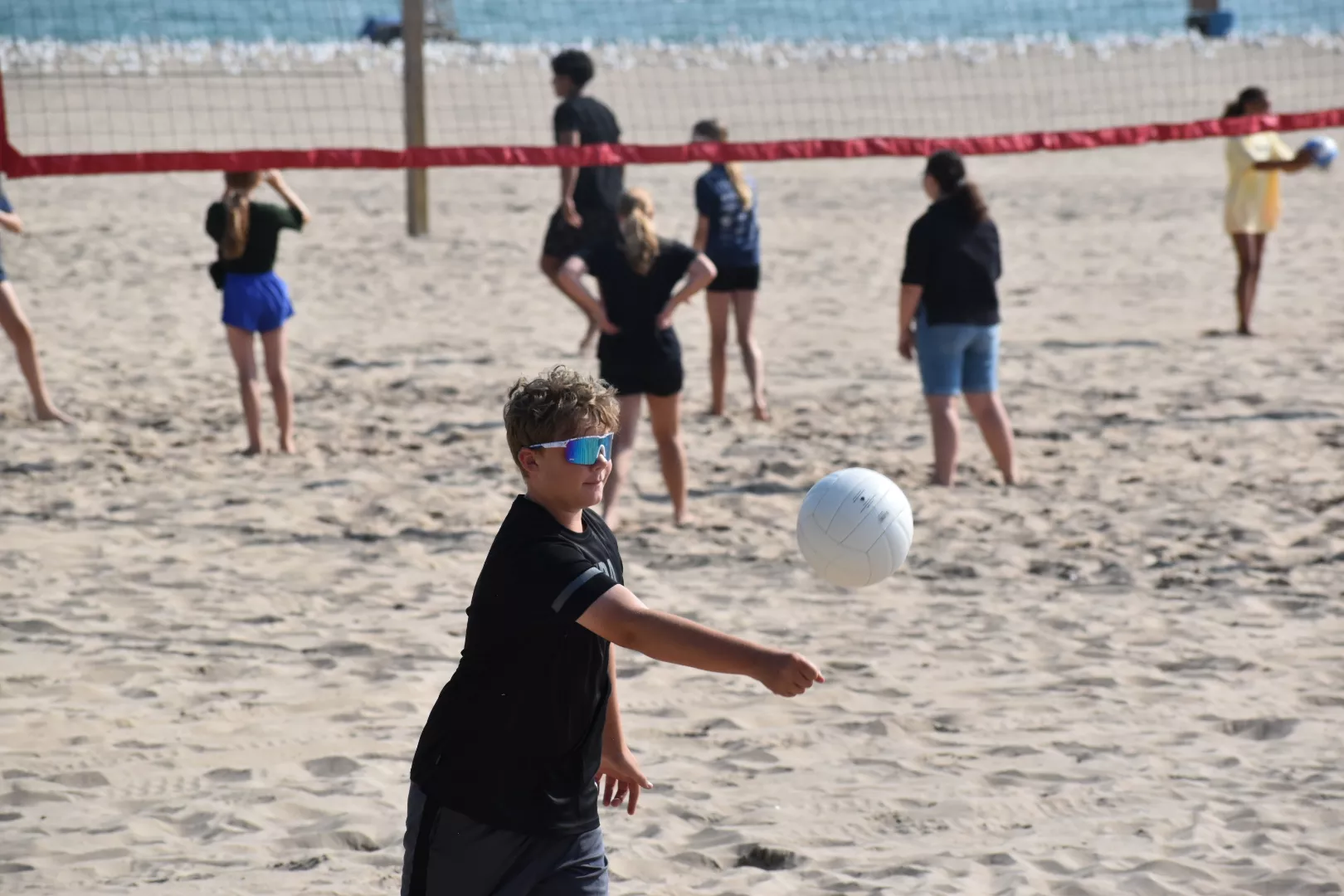 Teenager with sunglasses bumps volleyball on beach; net and other players in background.