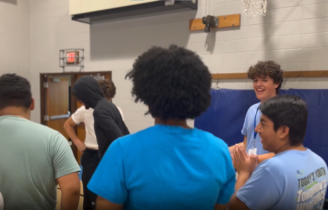 Group of diverse teenagers in a gymnasium, some wearing matching blue shirts with a youth program logo.