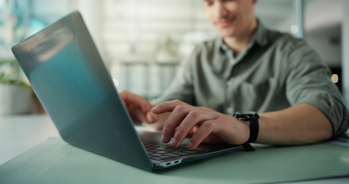 Person types on a laptop, smiling slightly. Laptop is on a desk with a plant in the background.