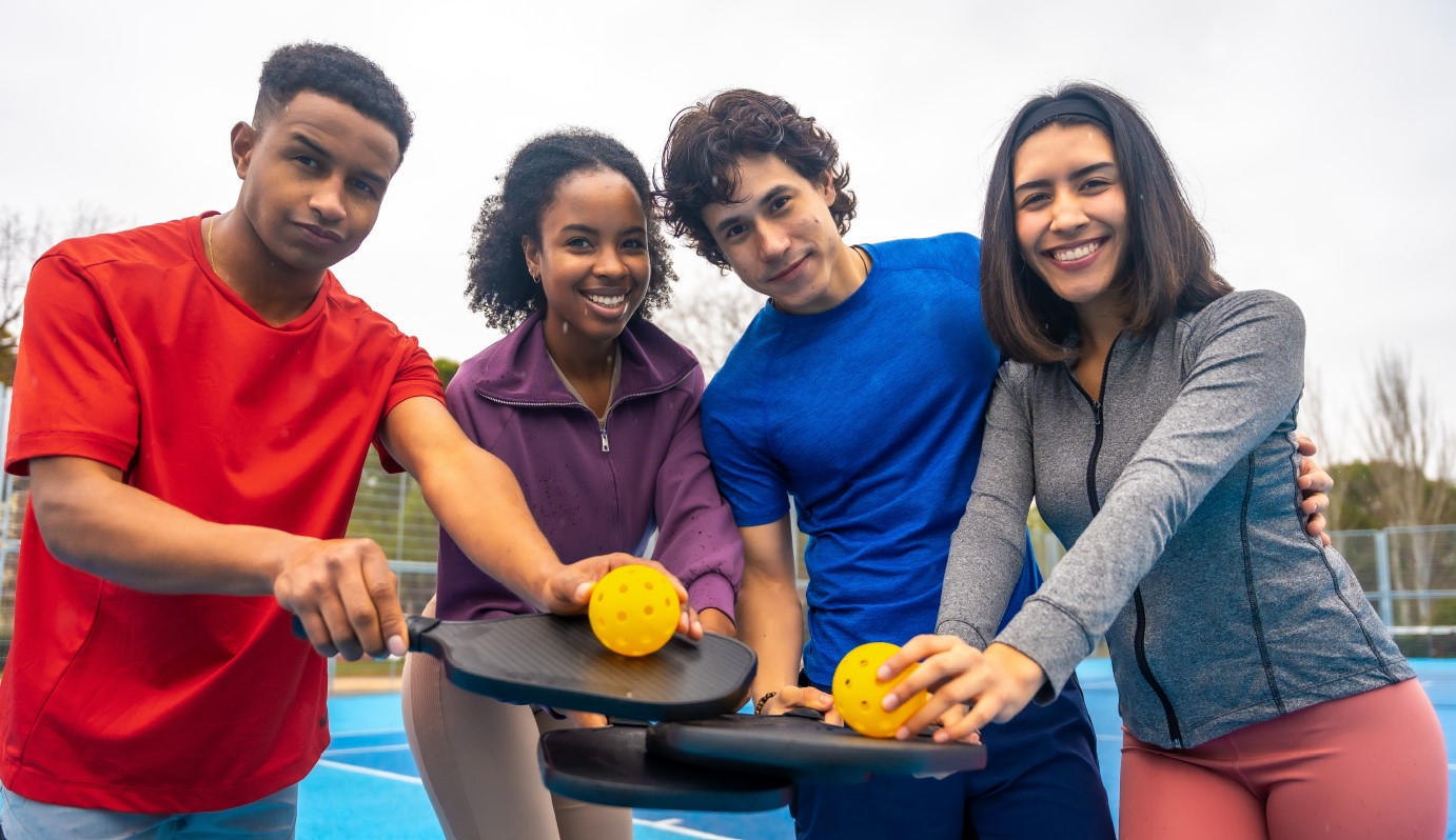 Four smiling young adults with paddles and pickleballs on a blue court, promoting pickleball.