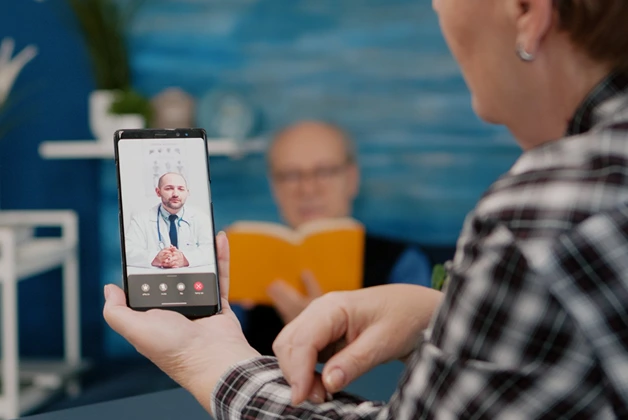 Senior woman using smartphone for telehealth consultation with a male doctor. Man reading in background.