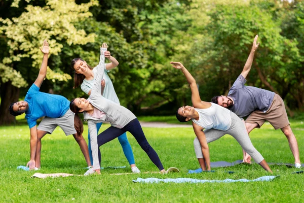 Group of diverse people doing yoga outdoors on mats in a park, triangle pose with arms raised.