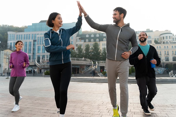 Group of four people jogging outdoors, woman giving high-five to man. Buildings in background.