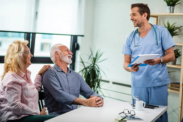 Smiling doctor in scrubs reviews chart with elderly couple at table in a well-lit room.